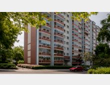 Mehrstöckiges Wohnhaus mit einer Fassade aus verglasten Balkonen mit roten und weißen Elementen, umgeben von grünen Bäumen und Büschen. Im Vordergrund verläuft eine Straße mit einem geparkten roten Auto, angrenzend an den Gehweg.