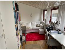 Kleinraum-Büro oder Wohnbereich mit Holzboden und holzverkleideter Decke. Ein weißes Sofa mit Kissen und Hocker steht vor großen Fenstern mit weißen Vorhängen, daneben ein schlichter Schreibtisch mit Stuhl und Arbeitsmaterialien; links ein Regal mit Büchern. Ein roter Teppich liegt zentral im Raum.