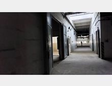 Ein langer, dunkler Flur in einem industriellen Gebäude mit hohen Decken und sichtbaren Rohren. Mehrere offene Metalltüren führen in angrenzende Räume, am Ende des Flurs ist eine Person vor großen Fenstern zu sehen.