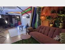 Ein kreatives Fotostudio mit einer gemütlichen Sitzecke, bestehend aus einem rosa Sofa, grünen Pflanzen und einer Stehlampe. Im Hintergrund befinden sich farbige Hintergrundrollen, Studiobeleuchtung und moderne Hocker vor einer weißen Fotofläche.