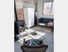 Gemütlicher Raum im Industriestil mit freigelegter Backsteinwand, einem schwarzen Holzofen, einem grauen Sofa auf einem Sisalteppich, einem kleinen Beistelltisch und einem Sessel im Vordergrund. Große Fenster lassen viel Tageslicht herein, daneben stehen weiße Metallschränke.