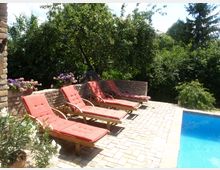 Gartenbereich mit vier roten Liegestühlen auf einem gepflasterten Boden neben einem Pool, im Hintergrund eine niedrige Ziegelmauer, blühende Blumen und dichte grüne Bäume.