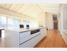 Moderne, helle Küche mit einer weißen Kücheninsel, ausgestattet mit einem Waschbecken und einer Kochplatte. Der Raum hat einen Holzboden, hohe Decken mit sichtbaren Balken, sowie große Fenster, die viel Tageslicht hereinlassen und einen Blick auf die Außenwelt bieten.