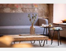 Ein gemütliches Wohnzimmer mit einer grauen Couch im Hintergrund, einem niedrigen Holztisch mit Metallbeinen im Vordergrund und zwei runden Beistelltischen, auf denen Kerzen und Glasgefäße stehen. Der Raum hat eine rustikale Wand mit sichtbaren Putzstellen und warmem Licht.