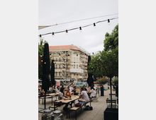 Außenbereich eines Cafés mit langen Holztischen und Bänken, umgeben von Bäumen und Dekorationslichtern, im Hintergrund ein saniertes Gebäude mit Gerüst und eine belebte Straße.