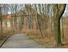 Gepflasterter Weg in einem parkähnlichen Bereich, umgeben von kahlen Bäumen und Sträuchern, mit einem mehrstöckigen, historischen Ziegelgebäude im Hintergrund.