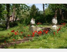 Grüne Parklandschaft mit roten und gelben Tulpen, zwei steinernen Zierurnen auf Sockeln und einer kleinen Treppe. Im Hintergrund stehen Bäume und ein Holzhaus ist teilweise sichtbar.