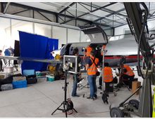 Innenansicht eines Hangars mit einem kleinen Flugzeug, das von mehreren Technikern in orangefarbenen Warnwesten für Film- oder Fotoaufnahmen vorbereitet wird. Im Vordergrund sind Kamerastative, Beleuchtungsgeräte und Kisten mit Ausrüstung sichtbar; im Hintergrund hängt ein großer blauer Stoffvorhang.