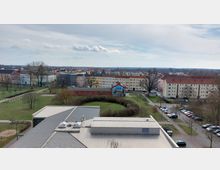 Blick auf eine städtische Wohngegend mit mehrstöckigen Gebäuden in verschiedenen Farben, umgeben von Grünflächen und einem kleinen Park. Im Vordergrund befindet sich ein modernes Flachdachgebäude und ein Parkplatz, im Hintergrund erstreckt sich eine flache Landschaft.