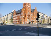 Straßenszene mit einem markanten, roten Backsteingebäude mit Türmen und vielen Fenstern im neugotischen Stil, daneben ein modernes Gebäude mit großen Glas- und Steinfassaden. Im Vordergrund eine breite Kreuzung mit Straßenmarkierungen, Ampeln und geparkten Autos.