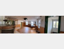 Ein schlicht eingerichteter Raum mit Holzboden, holzvertäfelten Wänden und mehreren Fenstern, die Tageslicht hereinlassen. Im Raum befinden sich Tische, Stühle und ein Projektor an der Decke, während im Vordergrund ein alter Kamin sichtbar ist.