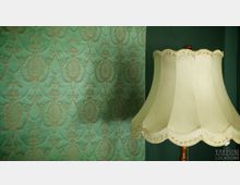 Grüne Wand mit ornamentaler, goldfarbener Musterung im Barockstil, daneben ein beigefarbener Lampenschirm mit welligem Rand und feinen Verzierungen.