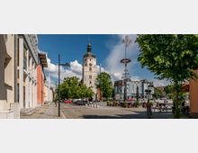 Straßenszene in einer historischen Kleinstadt mit einer gepflasterten Fußgängerzone, flankiert von bunten Gebäuden auf der linken Seite. Im Hintergrund steht ein markanter Kirchturm mit einer Uhr, umgeben von Bäumen und einem Maibaum auf einem zentralen Platz.