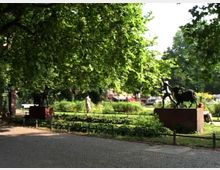 Ein Park mit viel Grünfläche, großen Bäumen und mehreren Skulpturen, darunter eine Statue auf einem Backsteinsockel, die eine Reiterfigur darstellt. Im Hintergrund sind Spazierwege, eine Wiese und Gebäude zu erkennen.
