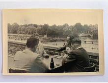 Schwarz-weiß-Fotografie von vier Männern, die an einem Tisch im Freien Karten spielen. Im Hintergrund ist ein von Bäumen umgebener Fluss oder See mit einer Holzbrücke und einem Steg zu sehen.