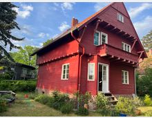 Rotes Holzhaus mit Satteldach und weiß umrahmten Fenstern, teilweise überdachtem Balkon und Ziegelsockel, umgeben von einem Garten mit Pflanzen und Rasen. Im Hintergrund sind Bäume und ein weiteres Gebäude sichtbar.