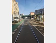 Straßenansicht einer städtischen Straße mit Straßenbahnschienen in der Mitte, einem Fahrradweg auf der linken Seite und einem Fußgängerweg rechts. Im Hintergrund führt eine grüne Stahlbrücke für Bahnschienen über die Straße, umgeben von mehrstöckigen Wohn- und Geschäftsgebäuden.