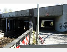 Ein Bahngleis unter einer Betonbrücke, daneben ein gepflasterter Gehweg, der ebenfalls unter die Brücke führt. Die Wände der Brücke sind mit Graffiti bedeckt, und ein rotes Fahrrad ist an einem Laternenpfahl angelehnt.