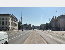 Breite Straße in einer städtischen Umgebung mit historischen Gebäuden auf beiden Seiten, darunter ein neoklassizistisches Gebäude rechts und ein barockes Gebäude links. Im Hintergrund sind der Berliner Fernsehturm und die Kuppel des Berliner Doms sichtbar.