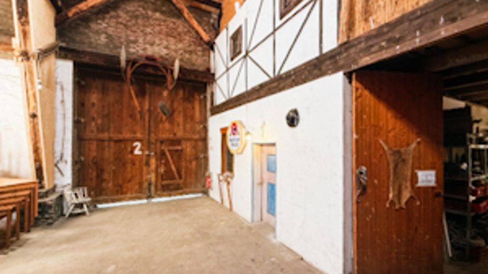 Innenraum einer rustikalen Scheune oder Lagerhalle mit hohen Holzbalken, sichtbaren Dachträgern und teils weiß verputzten Wänden. An den Wänden hängen landwirtschaftliche Geräte und Dekorationen; rechts führt eine offene Holztür in einen Nebenbereich.