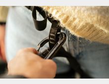 Nahaufnahme von schwarzen Karabinern und Gurtbändern eines Klettergurts, die an einer Person mit Jeans und einem beigen Strickpullover befestigt sind. Der Hintergrund ist unscharf und zeigt keine klar erkennbaren Umgebungselemente.