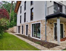 Mehrstöckiges Wohngebäude mit heller Putzfassade und Natursteinverkleidung im unteren Bereich. Große Fenster und Türen blicken auf einen gepflegten Garten mit einem schmalen gepflasterten Weg, der von Pflanzen und Rasengras umgeben ist.