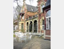 Historische Villa mit roter Ziegelfassade und dekorativen Steinverzierungen, großen Rundbogenfenstern und einer Terrasse mit weißen Metallstühlen und Tischen; umgeben von Bäumen und einem Gartenweg.