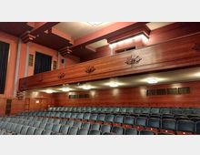 Innenraum eines kleinen Theatersaals mit einer hölzernen Empore und Reihen von gepolsterten Kinositzen. Die Wände sind mit Holzpaneelen verkleidet, und die Decke ist mit rechteckigen Deckenleuchten beleuchtet.