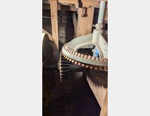 Blick in den Innenraum eines alten Holzgebäudes mit dicken Holzbalken und einer gemauerten Wand im Hintergrund. Im Vordergrund ist ein großes, teilweise verrostetes Zahnradgetriebe aus Metall mit Holz-Zahnkranz und einer senkrechten Welle zu sehen.