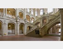 Historische Innenansicht eines prunkvollen Treppenhauses mit symmetrischen, geschwungenen Steintreppen, verzierten Balustraden und hohen Rundbögen. Die Architektur zeigt reichhaltige Steinornamente, hohe Fenster und Säulen in einem hellen, von Tageslicht erfüllten Raum.