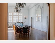 Eleganter Speisesaal mit einer langen gedeckten Tafel, dekoriert mit blauen Vasen und Trockenblumen. Der Raum ist lichtdurchflutet, hat Holzparkett, weiße Wände mit Stuckdetails, einen großen Spiegel sowie Kronleuchter und hohe Sprossenfenster.