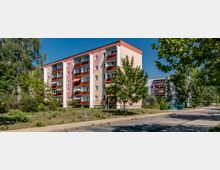 Mehrgeschossiges Wohngebäude mit farbiger Fassade in Rosa- und Beigetönen, roten Balkonen und weißen Fensterrahmen. Im Vordergrund befinden sich eine begrünte Fläche, Fußweg und einige Bäume, im Hintergrund weitere Gebäude und Müllcontainer.