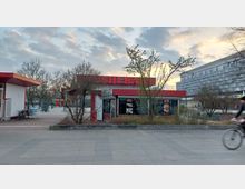 Ein kleiner Pavillon mit der Aufschrift „Sushi Bar“ und roten Akzenten, umgeben von kahlen Bäumen und Sträuchern, steht auf einem gepflasterten Platz. Im Hintergrund sind ein moderner mehrstöckiger Gebäudekomplex und ein Fahrradfahrer erkennbar.