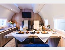 Innenraum einer luxuriösen Flugzeugkabine mit zwei schwarzen Ledersesseln, dekoriert mit Kissen und weißen Decken. Ein Holztisch ist mit feinen Speisen auf weißen Tischdecken gedeckt, und an den Seiten befinden sich kleine Fenster sowie Blumendekorationen auf den Ablageflächen.