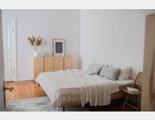Minimalistisch eingerichtetes Schlafzimmer mit einem Holzbett, weißer Bettwäsche und grauen Kissen. Im Hintergrund steht eine holzfarbene Kommode mit Dekorationen wie Trockenblumen, Kerzen und einem gerahmten Bild, auf einem Holzboden mit hellem Teppich.