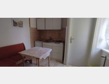 Kleine Küche mit einer weißen Küchenzeile, bestehend aus Hängeschränken, einer Spüle und einem Kochfeld. Im Vordergrund ein Esstisch mit Spitzen-Tischdecke und einer roten Sitzbank, daneben eine weiße Tür und ein Fenster mit Gardine.