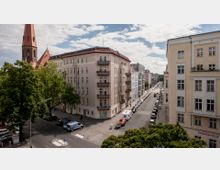 Straßenansicht in einer städtischen Umgebung mit klassischer europäischer Wohnarchitektur und einem spitzen Kirchturm im Hintergrund. Die Häuser haben helle Fassaden, Balkone, und die Straßen sind gesäumt von geparkten Autos und Bäumen.