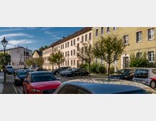 Straßenszene in einem Wohngebiet mit zweistöckigen, in hellen Farben gestrichenen Gebäuden, Bäumen entlang des Gehwegs und parkenden Autos auf Kopfsteinpflaster. Im Hintergrund ist eine Fassade mit Wandmalereien zu sehen.