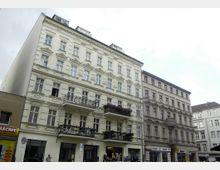 Mehrstöckiges Wohn- und Geschäftshaus im Stil des Historismus mit hellen Fassaden, verzierten Fensterrahmen und Balkonen, teilweise mit Blumen geschmückt; im Erdgeschoss befinden sich Ladengeschäfte. Rechts daneben ein ähnliches Gebäude mit schlichterer Gestaltung.