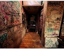 Schmaler Innenflur mit Holzfußboden, dessen Wände und Decke dicht mit Graffiti, Kritzeleien und Aufklebern bedeckt sind. Am Ende des Gangs stehen ein kleiner Tresen oder Automat und ein Feuerlöscher; rechts ist ein Heizkörper mit weiteren Markierungen zu sehen.