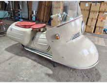 Innenraum einer Werkstatt oder Lagerhalle mit Betonboden, gestapelten Kartons und Holzplatten im Hintergrund. Im Vordergrund steht ein cremefarbenes Vintage-Kleinfahrzeug mit großer Frontverkleidung, Windschutzscheibe und zwei roten Sitzpolstern.