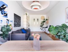 Modernes Wohnzimmer mit heller Einrichtung, einem grauen Sofa, Zimmerpflanzen und dekorativer moderner Kunst an den Wänden. Im Vordergrund steht ein Holztisch mit einer Vase, im Hintergrund sind weiße Türen und Regale sichtbar.