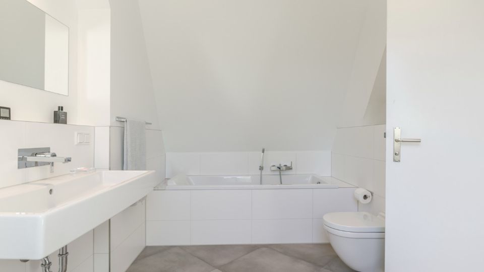 Helles, modernes Badezimmer mit schräger Dachdecke, eingebauter Badewanne in weißer Fliesenverkleidung und großformatigen grauen Bodenfliesen. Links ein breites Waschbecken mit Spiegel und Handtuchhalter, rechts ein wandhängendes WC neben einer offenen Tür.