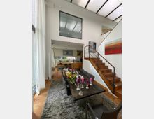 Moderne Innenaufnahme eines Wohn- und Essbereichs mit hohen Decken. Zu sehen sind ein großer Esstisch mit Kerzenleuchtern und Blumen, eine Treppe mit Holzstufen, eine offene Küche im Hintergrund und große Fenster mit weißen Vorhängen, die viel Tageslicht einlassen.