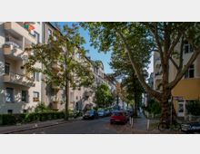 Straßenszene in einem Wohngebiet mit mehrstöckigen Gebäuden im Stil der 1950er-1960er Jahre, teilweise mit Balkonen. Die Straße ist von Bäumen gesäumt, mit geparkten Autos am Straßenrand und einem Straßenschild mit der Aufschrift „Badestraße“ an einer Ecke.