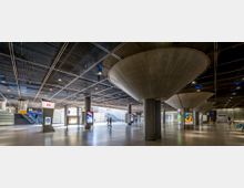 Geräumige Bahnhofshalle mit glatten Betonböden, auffälligen säulenartigen Betontrichtern, die bis zur Decke reichen, und einer Metall-Deckenverkleidung. Mehrere Werbebildschirme, Wegweiser und Zugänge zu Rolltreppen sind sichtbar.