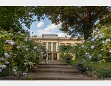 Historisches Gebäude mit klassizistischen Elementen, wie Säulen und großer Fensterfront, umgeben von einem Garten mit blühenden Pflanzen, Bäumen und dekorativen Steinvasen entlang einer kleinen Treppe.
