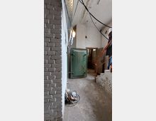 Ein schmaler, kellerartiger Raum mit weißen Backsteinwänden und einer Betondecke. Im Vordergrund steht eine grüne Metalltür mit Abnutzungsspuren, rechts führt eine Treppe mit einem roten Geländer nach oben, und Kabel hängen von der Decke.