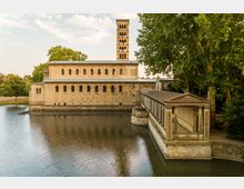Historische Kirche mit rechteckigem Hauptgebäude und einem markanten Glockenturm, umgeben von einer Wasserfläche. Rechts im Bild befindet sich ein überdachter Säulengang, der zu einem Eingang führt, umrahmt von Bäumen und Parklandschaft.