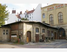 Backsteingebäude mit gelblichen Klinkern und mehreren hölzernen Türen, daneben ein moderneres Gebäude aus weißem Putz mit roten Ziegeldächern. Im Vordergrund ein gepflasterter Hof mit gelben Linien und ein roter Hydrant.